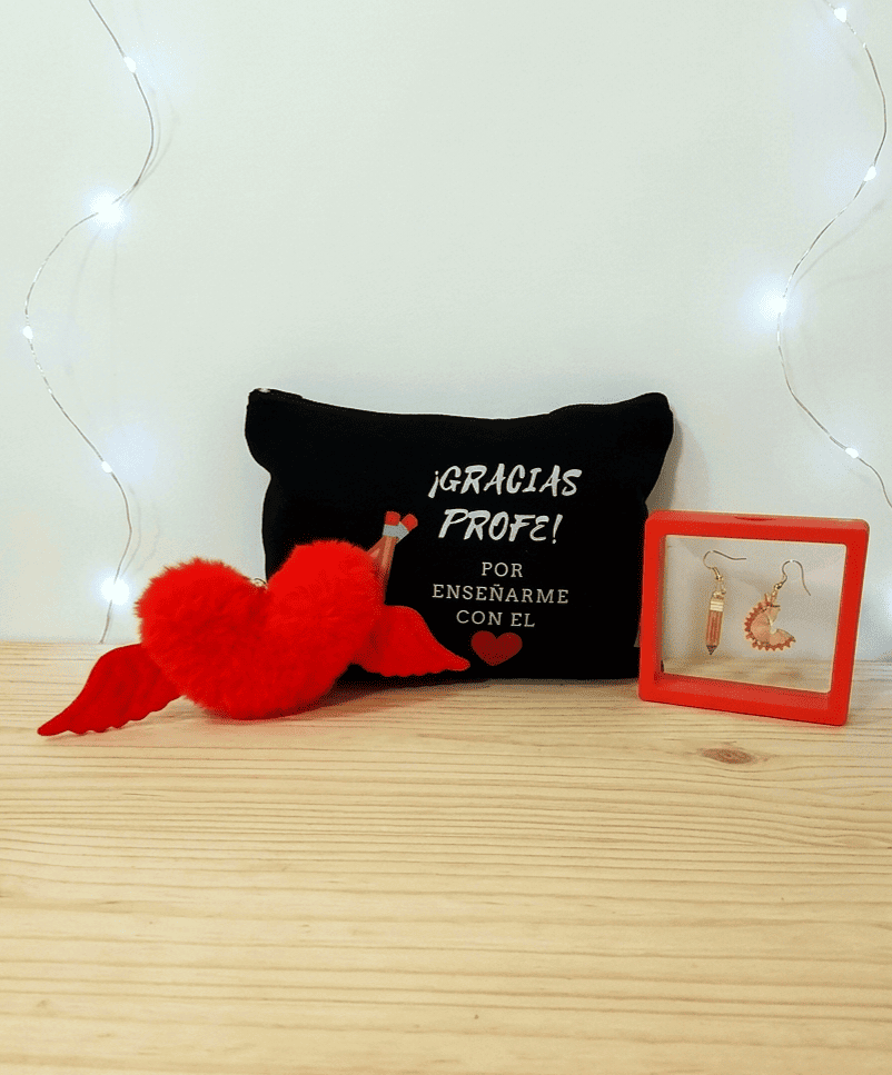 Conjunto de regalos del pack "Heart Gift Box"  formado por un llavero en forma de corazón en peluche rojo con alas,  en el medio puede verse un neceser negro con frase de agradecimiento para profesor en español con letras blancas y dibujo de un corazón rojo y dos lapiceros, por ultimo se puede ver unos pendientes uno de ellos en forma de lapiz y el otro de viruta de lapiz, presentados en caja de exhibición con marco rojo, todo sobre una superficie de madera clara. 