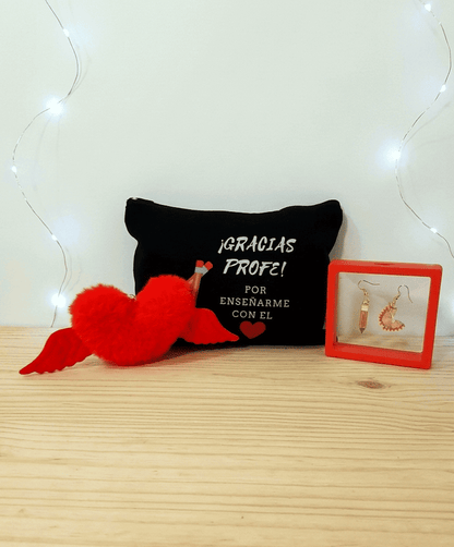 Conjunto de regalos del pack "Heart Gift Box"  formado por un llavero en forma de corazón en peluche rojo con alas,  en el medio puede verse un neceser negro con frase de agradecimiento para profesor en español con letras blancas y dibujo de un corazón rojo y dos lapiceros, por ultimo se puede ver unos pendientes uno de ellos en forma de lapiz y el otro de viruta de lapiz, presentados en caja de exhibición con marco rojo, todo sobre una superficie de madera clara. 