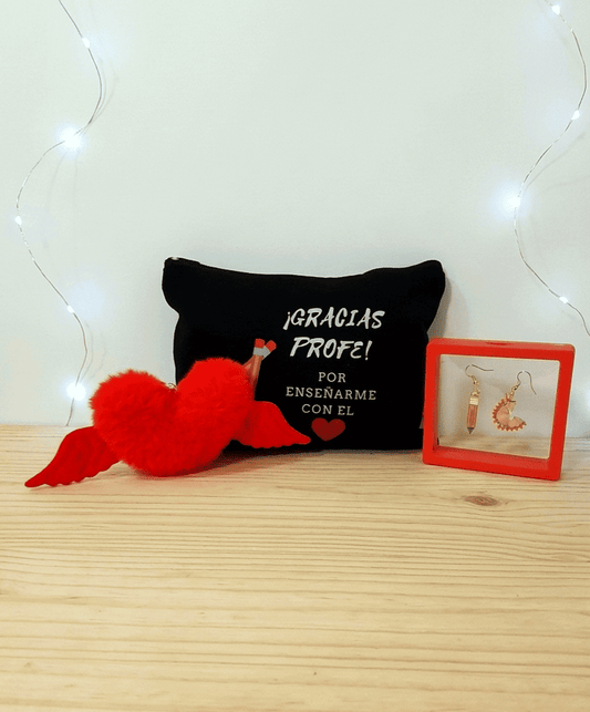 Conjunto de regalos del pack "Heart Gift Box"  formado por un llavero en forma de corazón en peluche rojo con alas,  en el medio puede verse un neceser negro con frase de agradecimiento para profesor en español con letras blancas y dibujo de un corazón rojo y dos lapiceros, por ultimo se puede ver unos pendientes uno de ellos en forma de lapiz y el otro de viruta de lapiz, presentados en caja de exhibición con marco rojo, todo sobre una superficie de madera clara. 