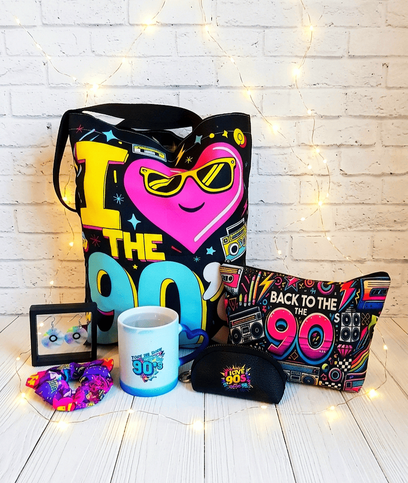 Conjunto de artículos y accesorios con diseño de los 90. Se pueden ver unos pendientes con forma de CD en caja de exibicion, un accesorio para el pelo con colores fluor, una taza termosensible con diseño de los 90, un monedero negro con el texto  " I Love 90's", un neceser con la frase "Back to the 90" en un fondo colorido, en el centro se puede ver una Bag Tote con dibujos grandes y coloridos. Todo el conjunto está sobre una superficie de madera blanca y pared blanca de ladrillo visto con luces led.