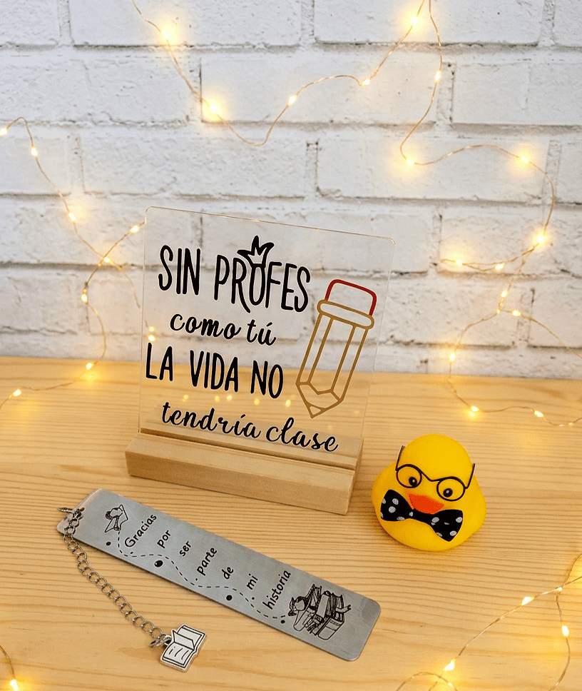 Primer plano de un conjunto de artículos de regalo para profes,  compuesto por una Placa Acrilica con la frase "Sin Profes como tu la vida no tendria clase", a su lado se encuentra un Patito de Goma amarillo con gafas y pajarita negra de lunares, por ultimo podemos ver un marcapaginas metálico con una frase de agradecimiento para Profes con detalles de motivos escolares con una cadenita de la que cuelga un dije con forma de libro abierto. Todo se encuentra sobre una superficie de madera clara y luces led.