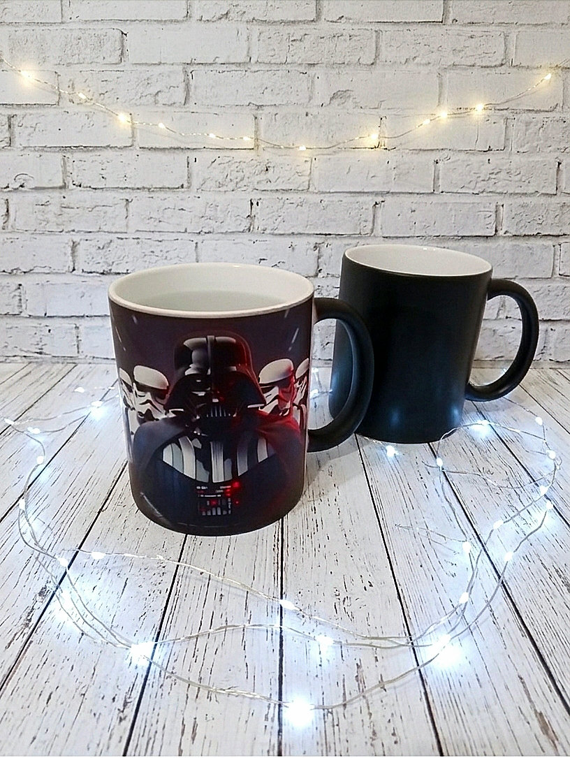 Una taza negra con diseño de Darth Vader, rodeada de luces led sobre suelo de madera blanca con una pared blanca con ladrillos de fondo.