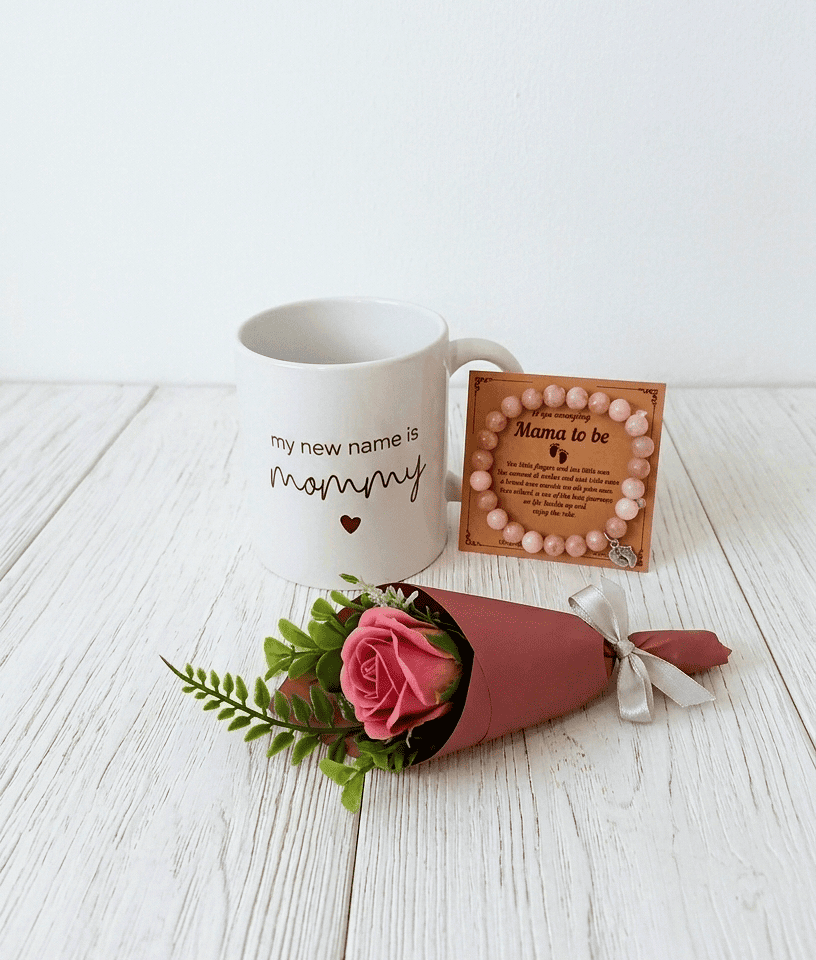 Conjunto de regalos compuesto por una taza blanca de ceramica con mensaje en inglés "My new name is mommy", pulsera de cuentas en colores pastel con dije de pies de bebé y una pequeña rosa eterna decorativa sobre superficie de madera blanca. 