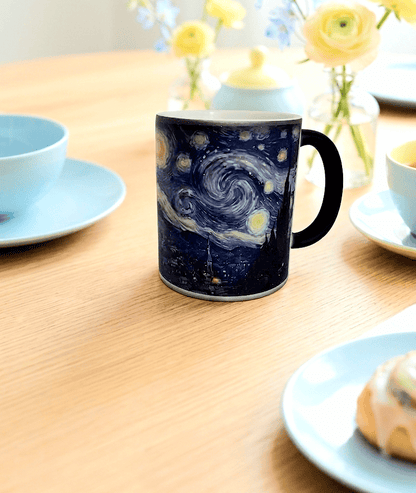 Taza de cerámica termosensible con bebida caliente en su interior donde aparece el diseño de el cuadro de "La noche estrellada" sobre una superficie de madera clara con un fondo de desayuno con accesorios y detalles en tonos azules y amarillos pastel.