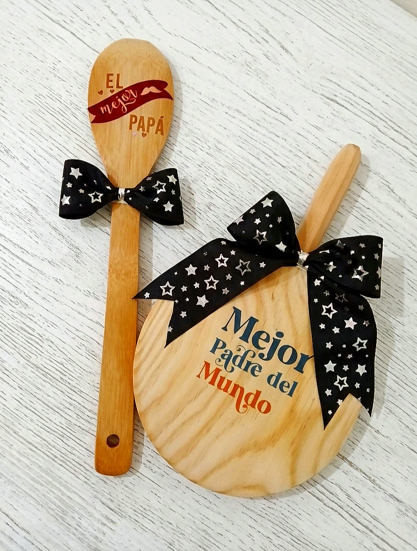 Vista superior de una cuchara de madera y una tabla de cocinar redonda en una mesa de madera blanca. La cuchara está grabada con  "El Mejor Papá " y la tabla tiene el texto "Mejor Padre del Mundo" Ambos objetos están decorados con lazos negros con estrellas plateadas