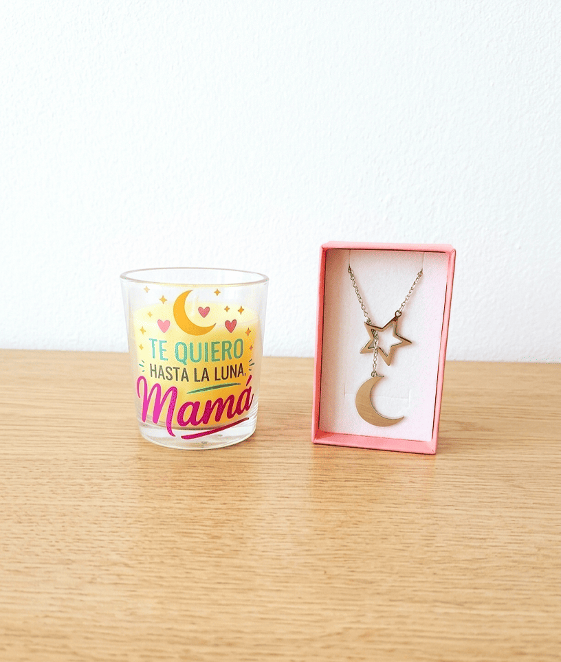 Vela en portavelas de cristal con la frase "Te quiero hasta la luna Mamá" con detalles de luna y estrellas junto a una caja rosa que contiene un colgante de Acero en color plateado compuesto por una estrella y media luna, todo sobre una superficie de madera clara. 