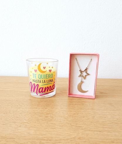 Vela en portavelas de cristal con la frase "Te quiero hasta la luna Mamá" con detalles de luna y estrellas junto a una caja rosa que contiene un colgante de Acero en color plateado compuesto por una estrella y media luna, todo sobre una superficie de madera clara. 