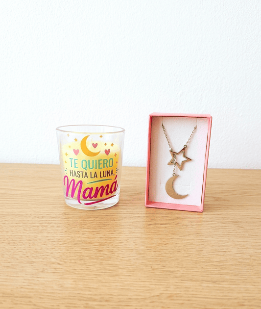 Vela en portavelas de cristal con la frase "Te quiero hasta la luna Mamá" con detalles de luna y estrellas junto a una caja rosa que contiene un colgante de Acero en color plateado compuesto por una estrella y media luna, todo sobre una superficie de madera clara. 