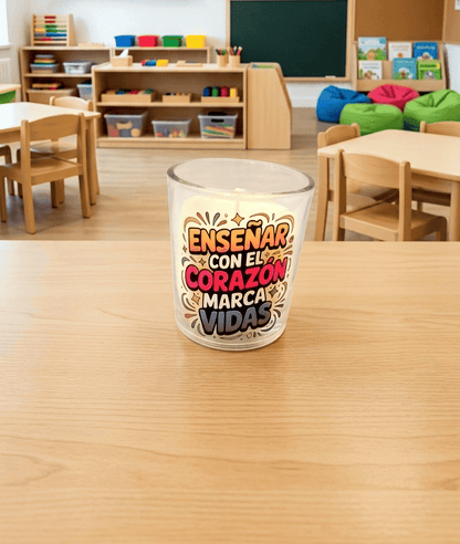 Primer plano de una vela en portavelas de cristal con la frase "ENSEÑAR CON EL CORAZÓN MARCA VIDAS" con tipografia moderna  de colores, se encuentra sobre una superficie de madera clara  y un fondo colorido de Escuela Infantil.