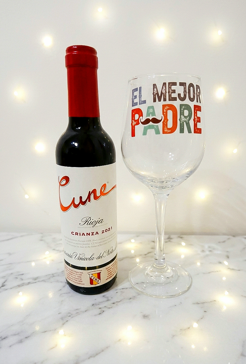 Vista frontal de un set de regalo que incluye una botella de vino Cune Rioja, una copa de vino personalizada con la frase " El Mejor Padre" en un fondo de mesa de marmol con luces led.